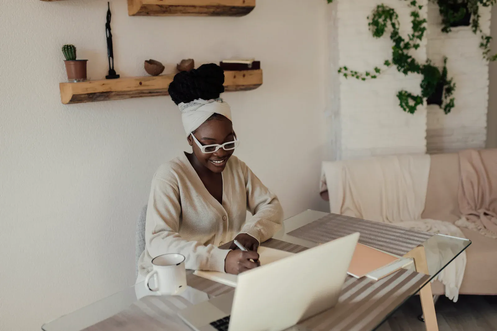 A woman writing a note beside her laptop in a warm, plant-filled home workspace