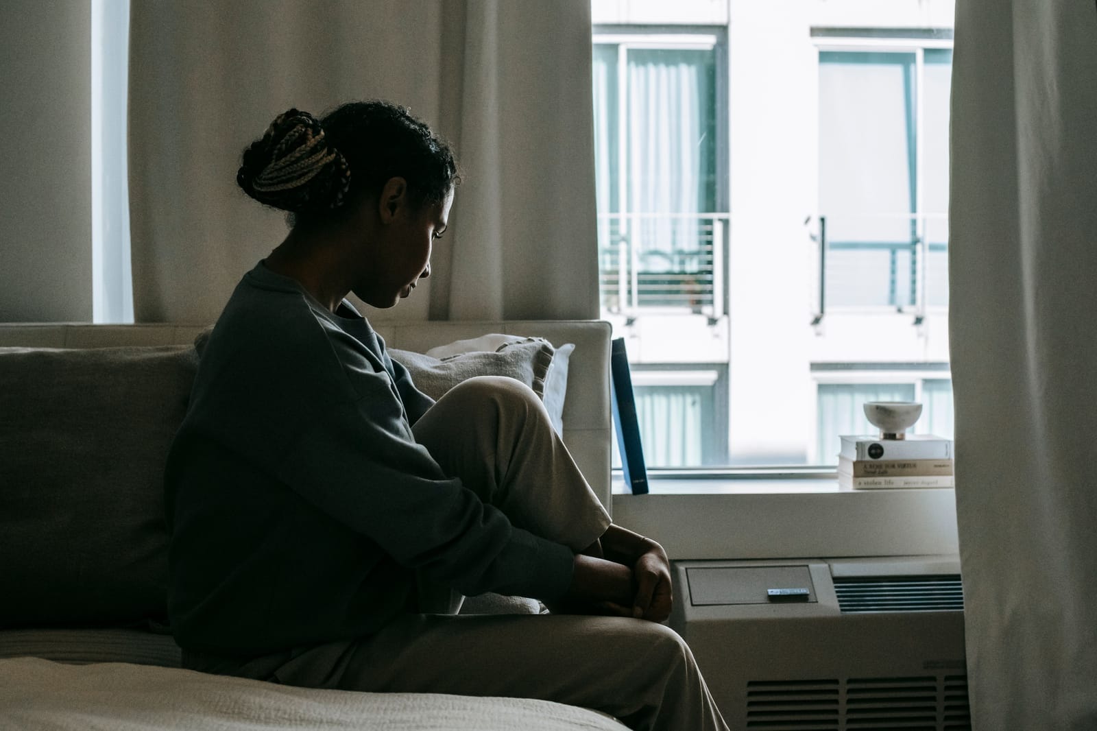 Black woman sitting quietly on a bed near a window at home, looking down in a reflective moment after a long day.