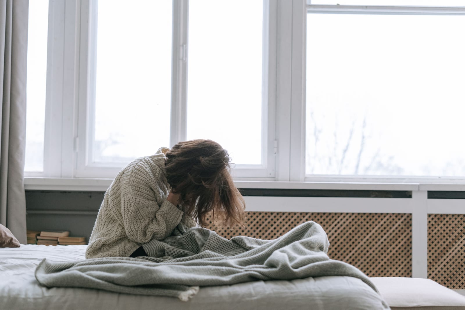White woman in a cream sweater sitting on a bed with her head lowered into her hands.